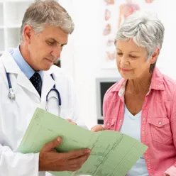 En lege i legefrakk viser en pasient noe som står skrevet på et ark.