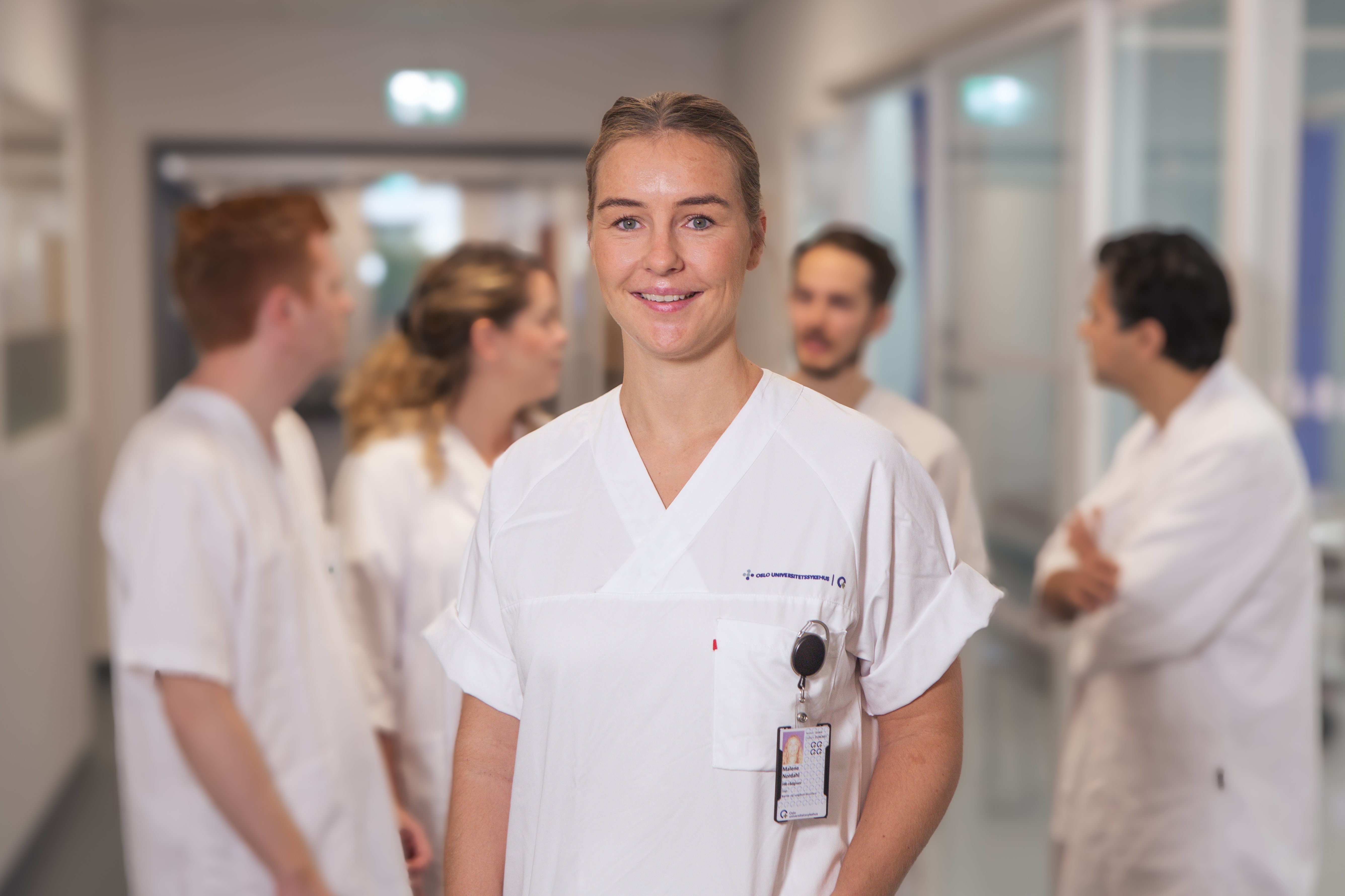 En helseansatt som står kledd i hvitt og smiler mot kameraet. Bak henne står det fire andre helsepersonell i hvitt og prater med hverandre.