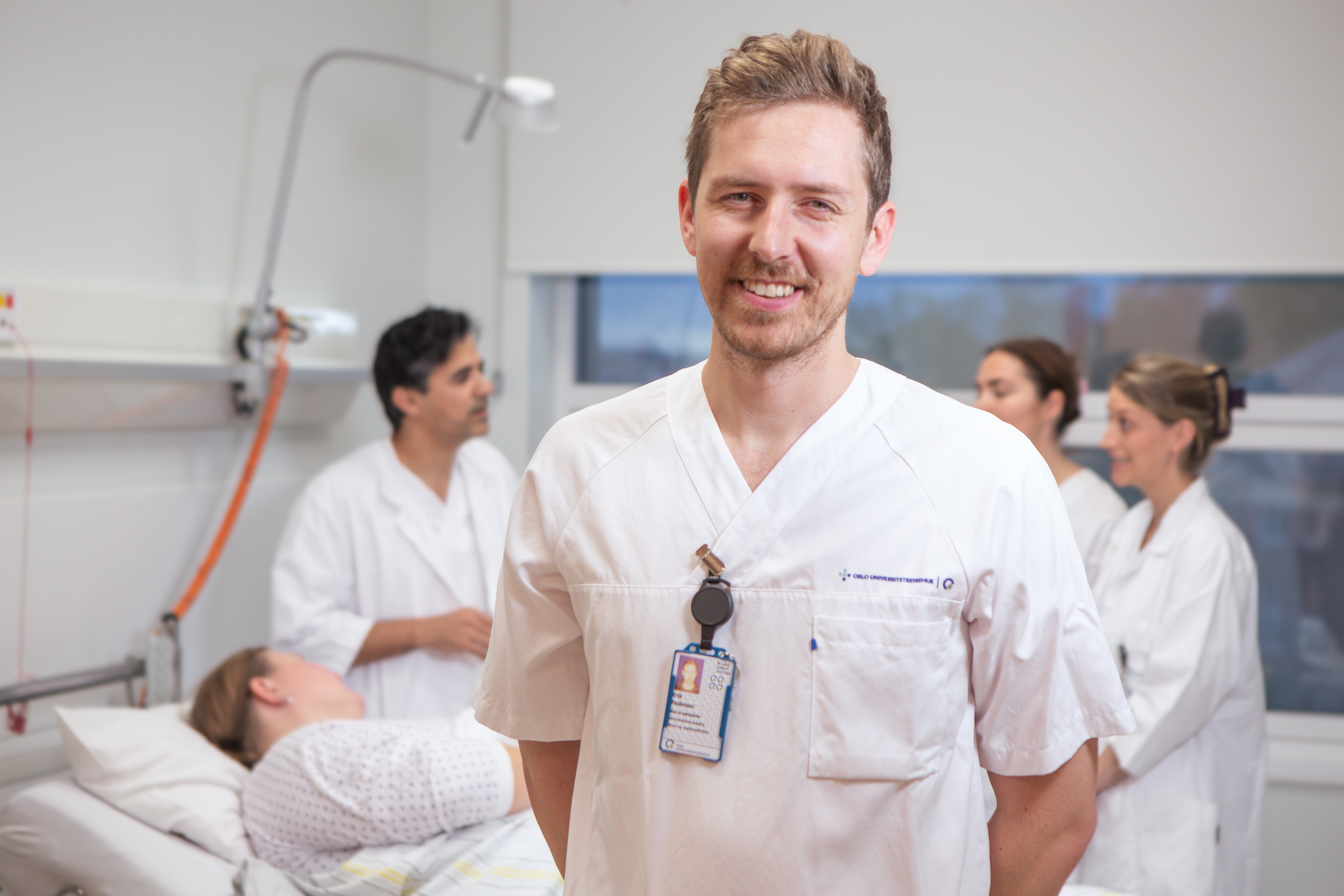 En sykepleier smiler mot kameraet. Bak ham står det tre andre helsepersonell rundt en pasient som ligger i en sykeseng.