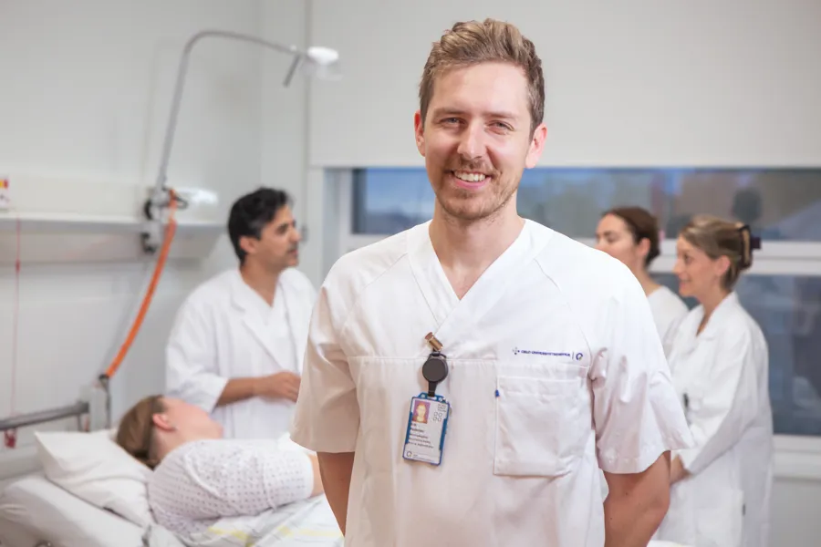 En sykepleier smiler mot kameraet. Bak ham står det tre andre helsepersonell rundt en pasient som ligger i en sykeseng.