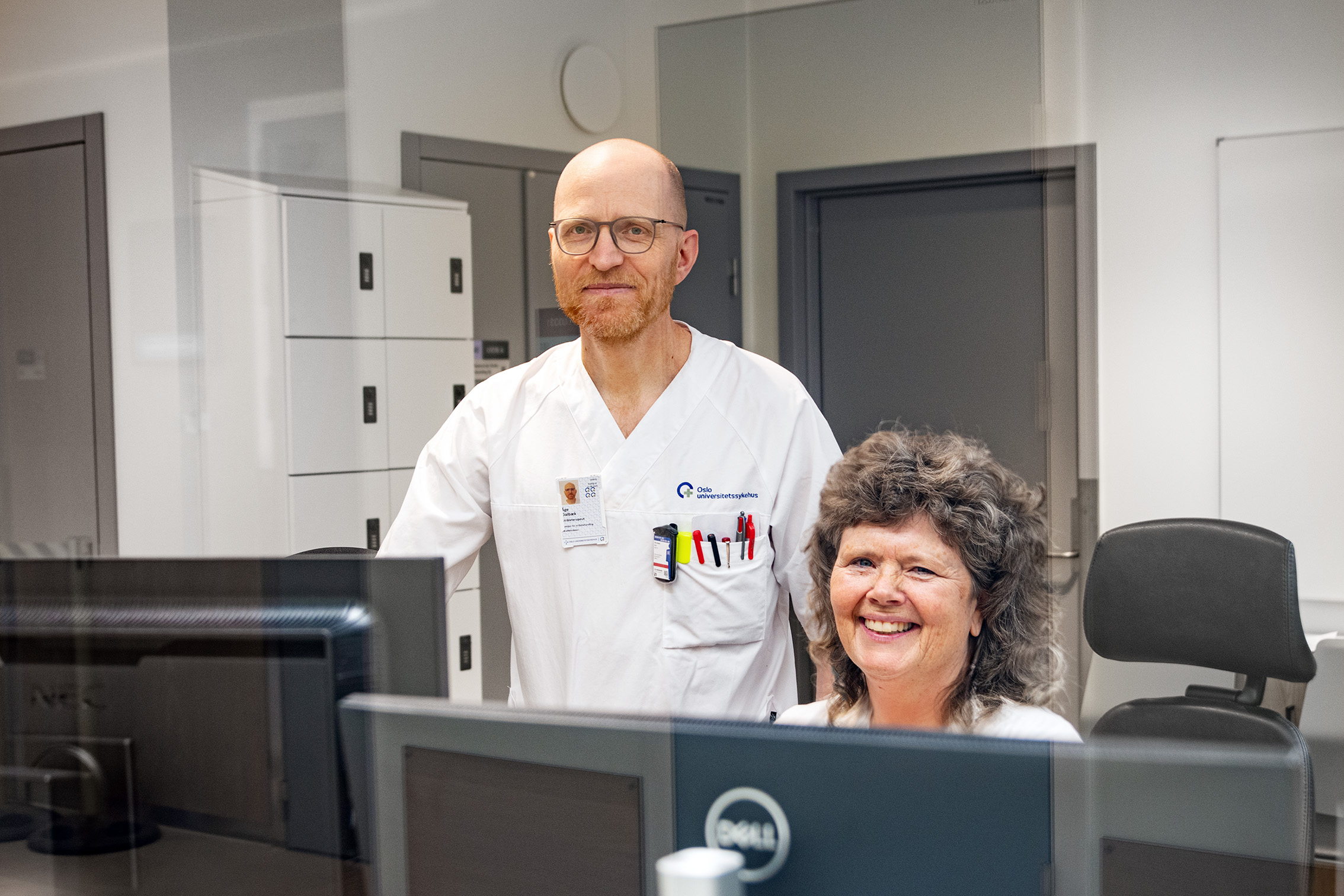 To ansatte sitter bak en pc og smiler inn i kamera