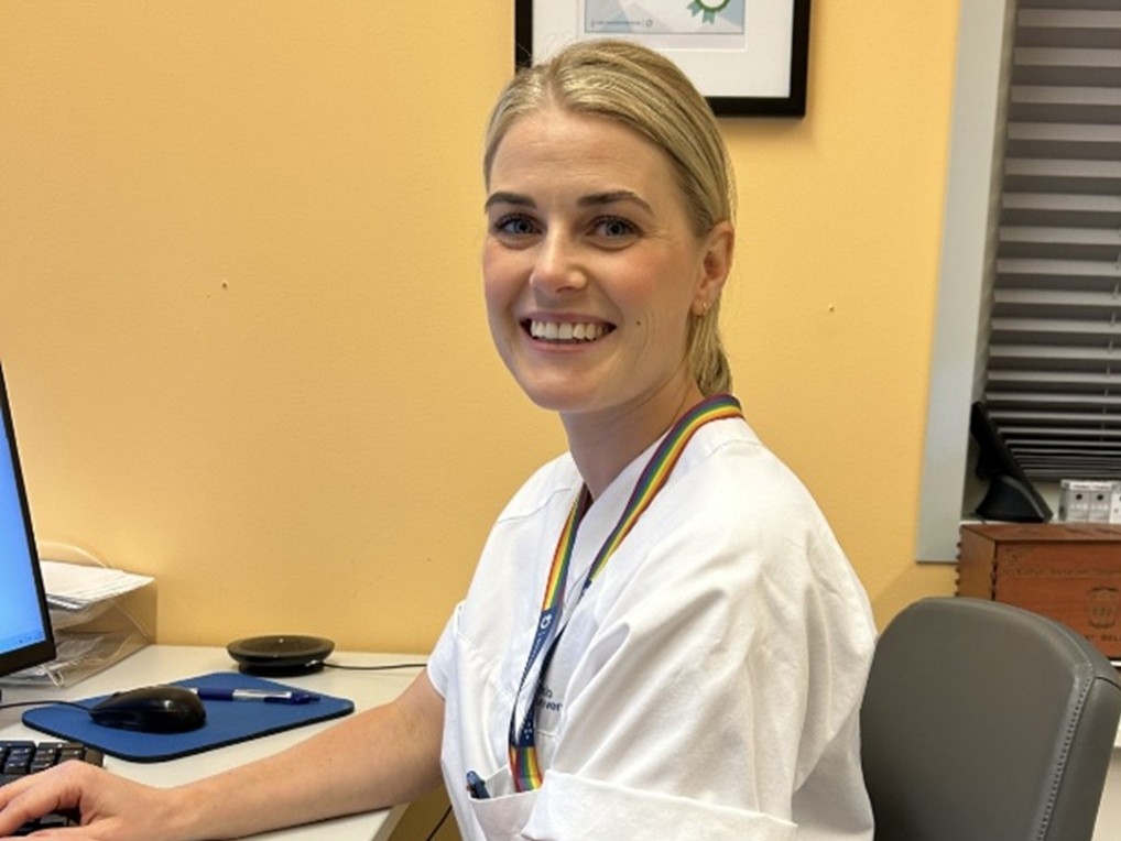 En ung kvinnelig helsefagarbeider i hvit uniform som sitter ved en pc og ser inn i kamera.