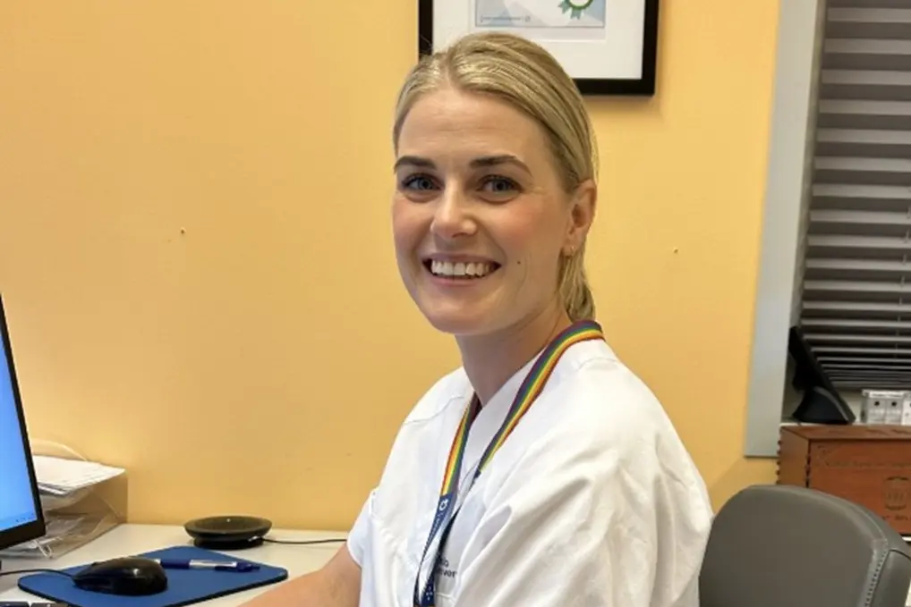 En ung kvinnelig helsefagarbeider i hvit uniform som sitter ved en pc og ser inn i kamera.