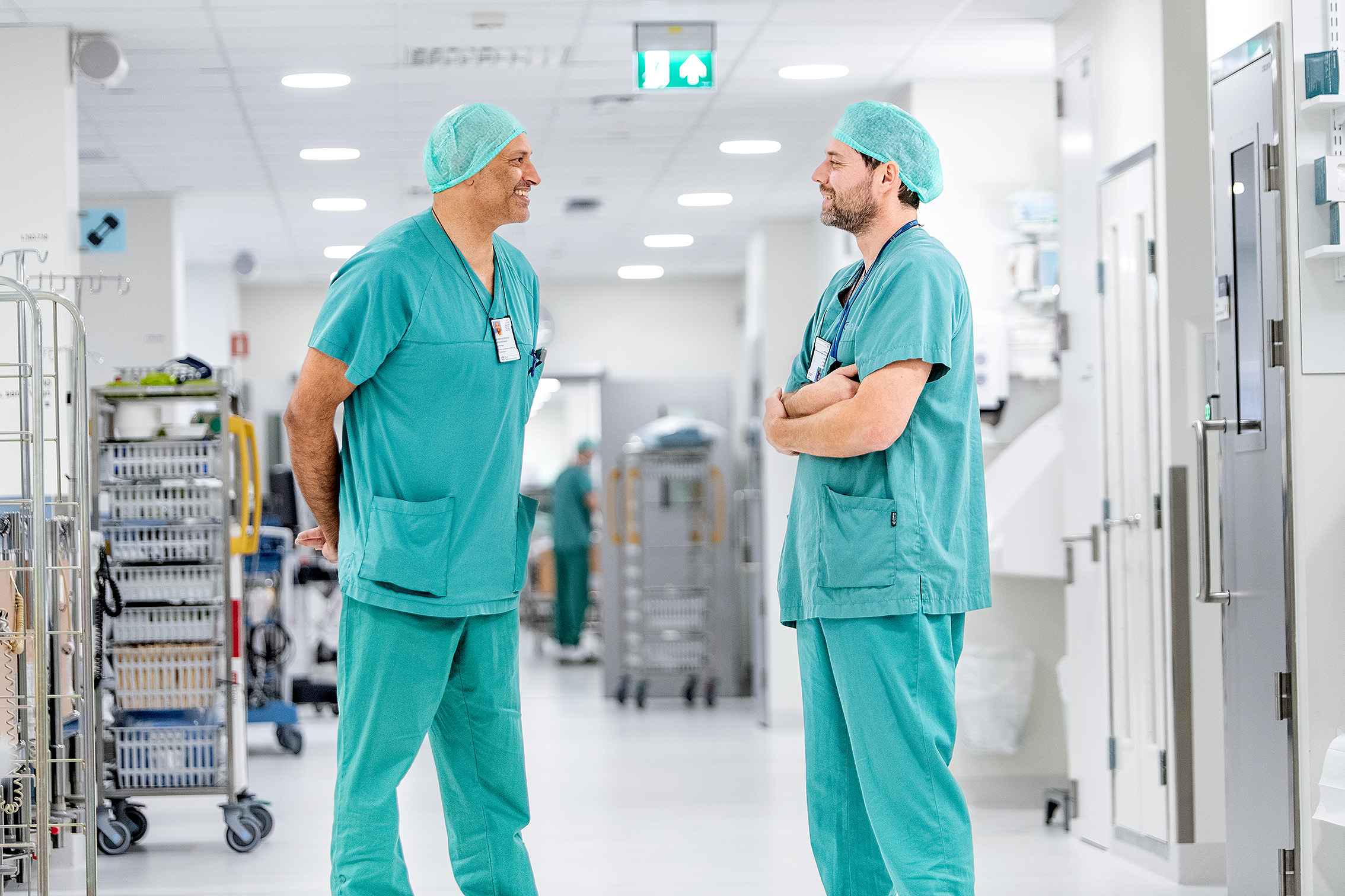 To kirurger ikledd grønt som står i en sykehuskorridor og prater sammen.