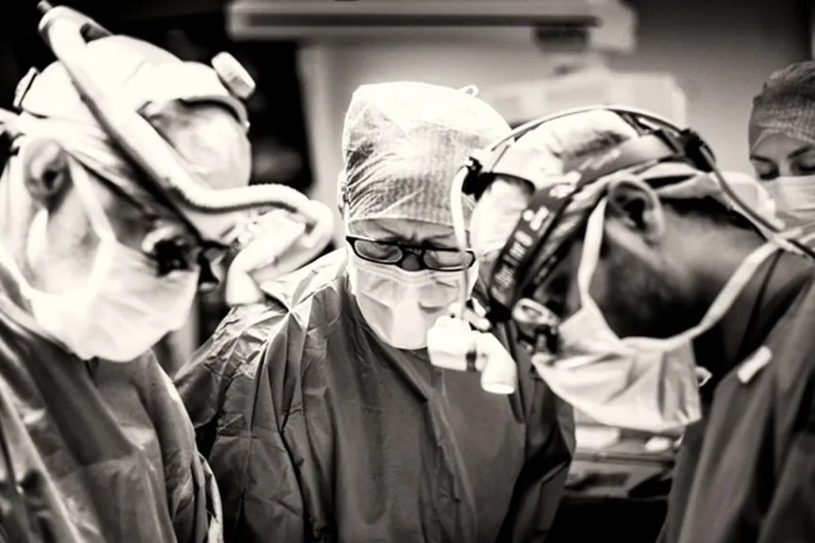 Tre kirurger står med munnbind, forstørrelsesbriller, hodeplagg og grønt antrekk.