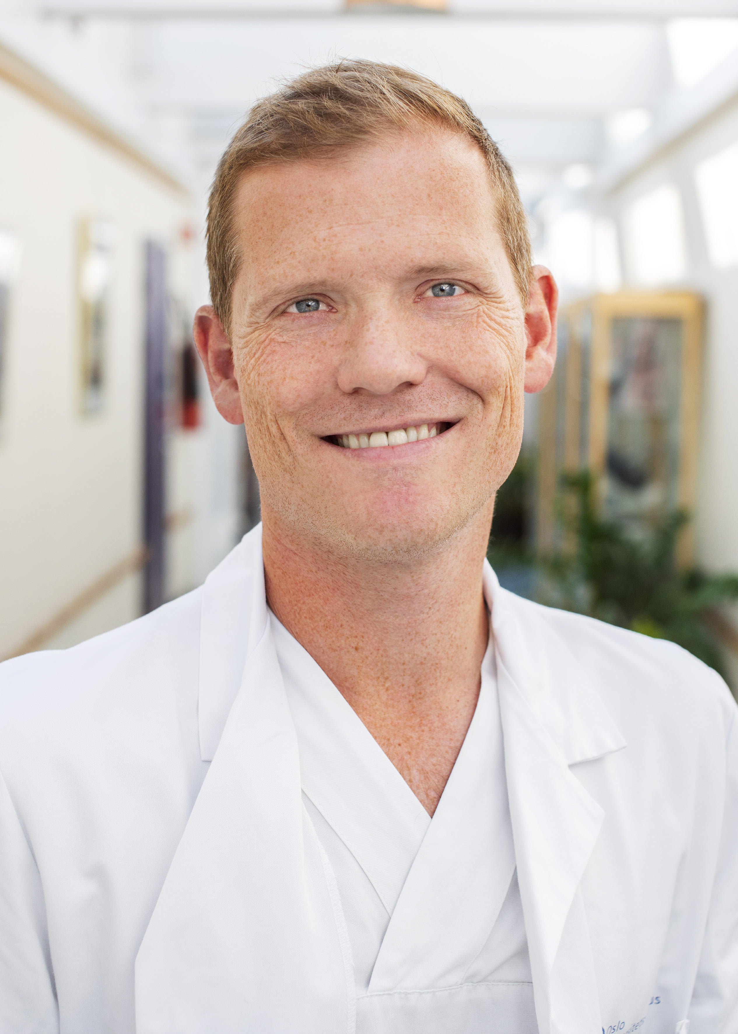 Portrettbilde av Jon Riise ikledd legefrakk. Han smiler mot kameraet.