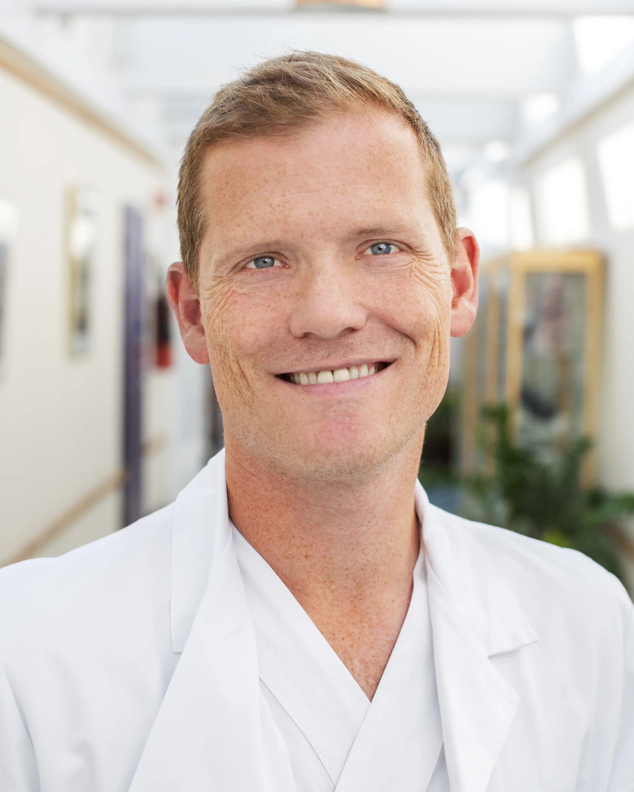 Portrettbilde av Jon Riise ikledd legefrakk. Han smiler mot kameraet.