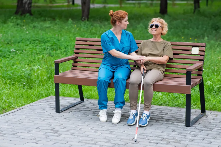 En eldre blind dame sittende på en benk med en sykepleier