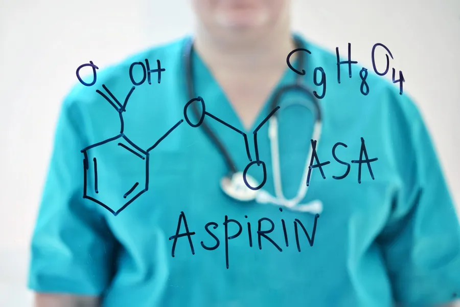 En gjennomsiktig tavle med den kjemiske formelen til acetylsalisylsyre og ordet Aspirin. En grønnkledd lege står i bakgrunnen.