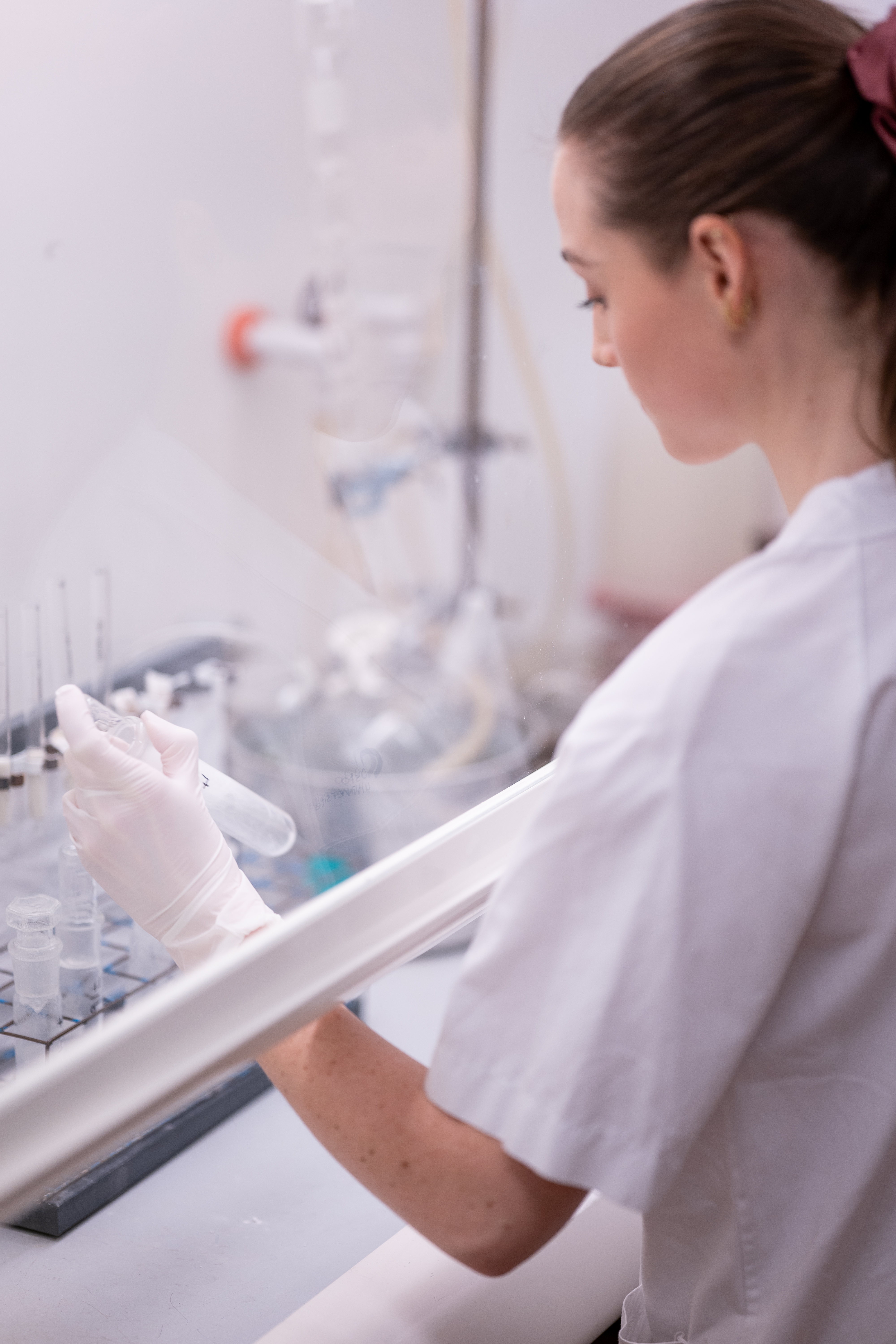 Laboratoriearbeider i sterilbenk holder et prøverør med klar væske, omgitt av annet analyseutstyr.