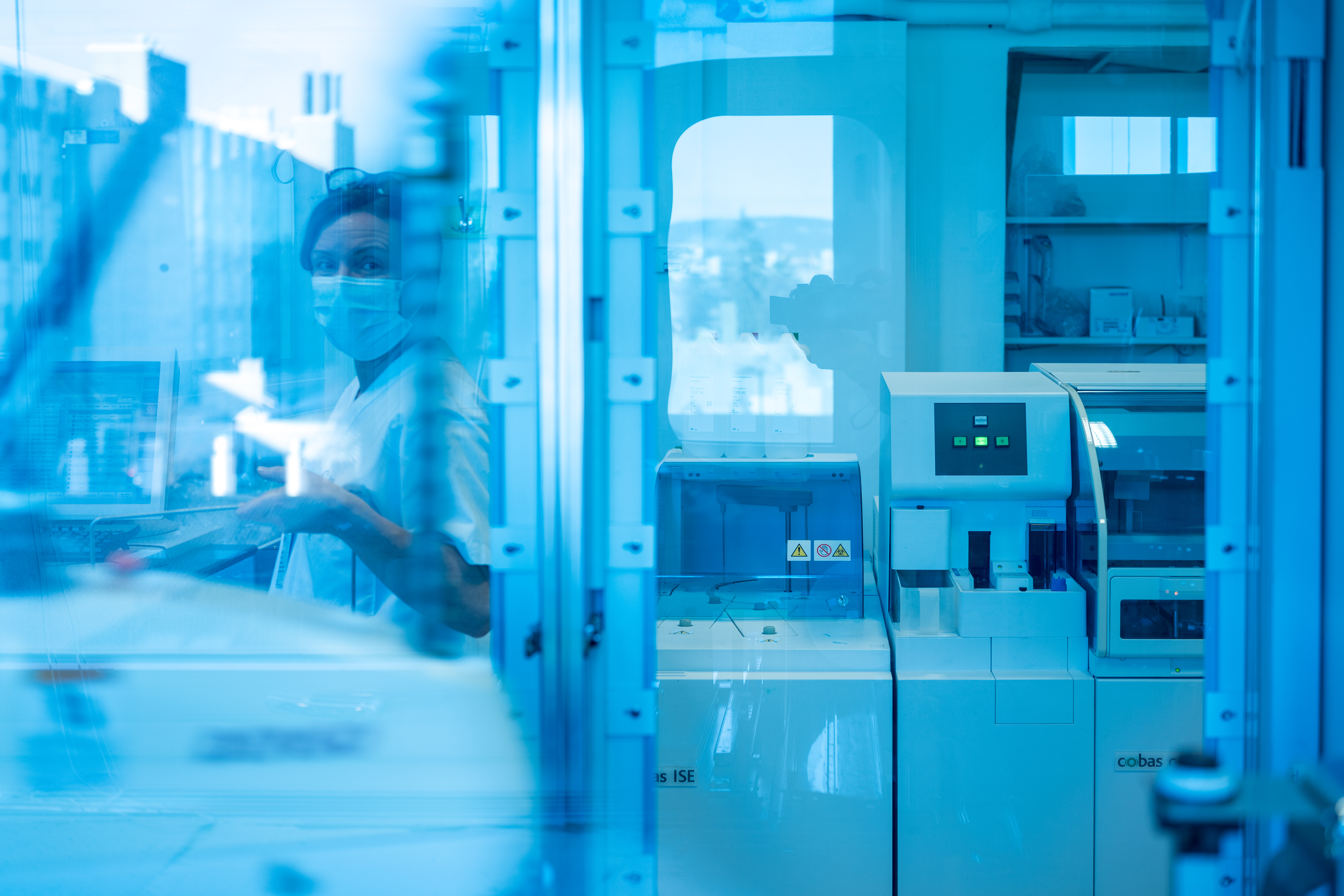 Laboratoriearbeider med munnbind sitter ved analyseutstyr og arbeider med prøver i et medisinsk laboratorium.