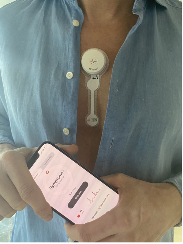 Plasteret med EKG-utstyr festet på brystkassen til en pasient. I hånden holder pasienten en mobiltelefon som viser hva EKG-plasteret har registrert.