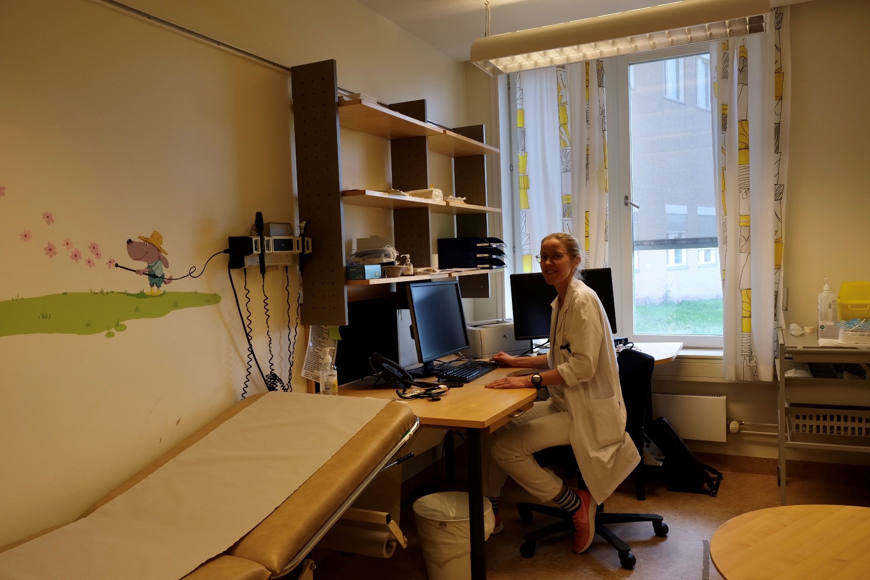 Et av konsultasjonsrommene. Her sitter en lege bak et skrivebord ved en datamaskin. Foran henne står et undersøkelsesbord.