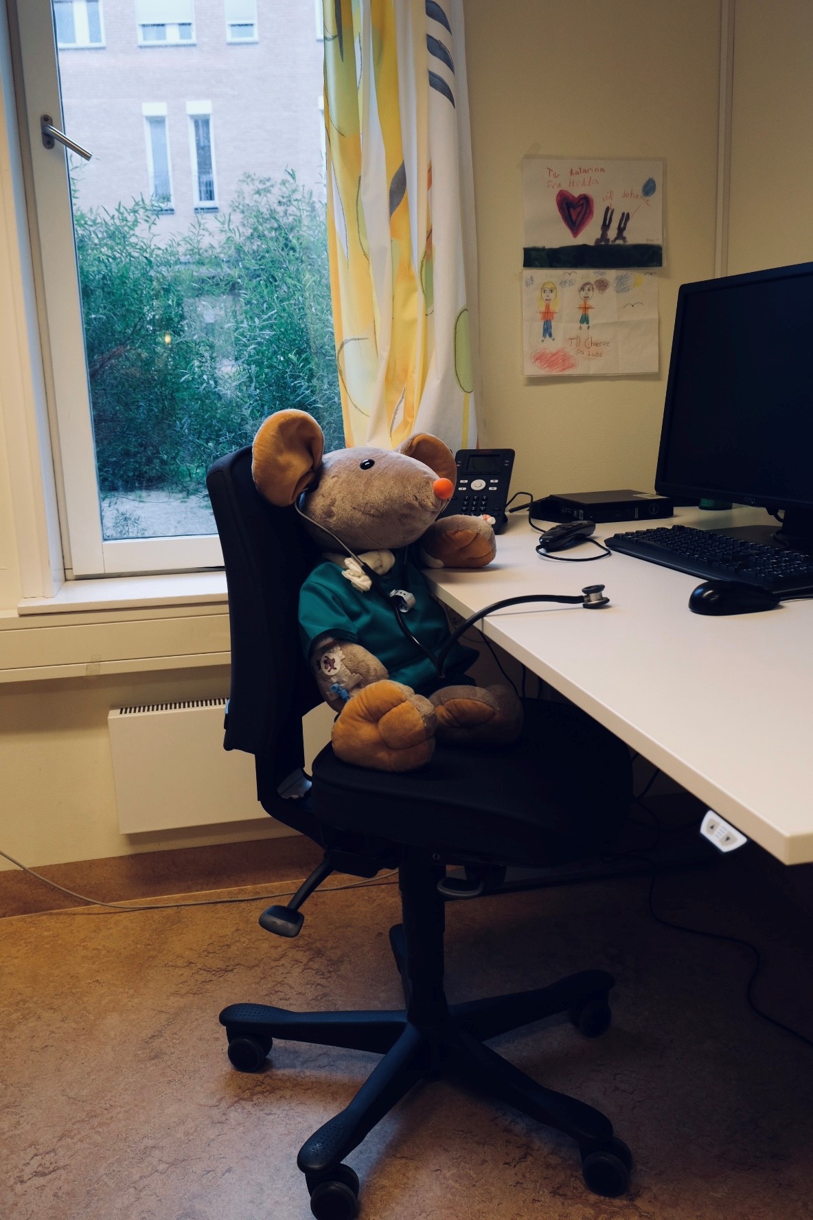 Sykehusmusa Rasmus sitter ved et skrivebord og lytter til bordet med et stetoskop.