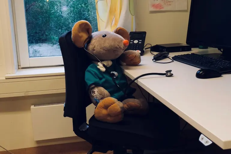 Sykehusmusa Rasmus sitter ved et skrivebord og lytter til bordet med et stetoskop.