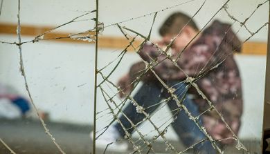 En ung gutt med hettejakke sitter med hodet bøyd bak knust glass