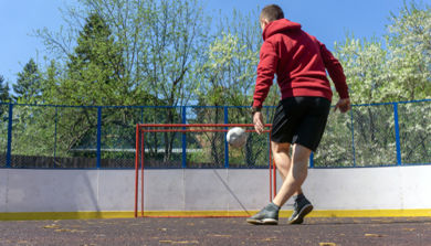 Ungdomsgutt med sort shorts og rød hettegenser sparker ball mot et fotballmål. 