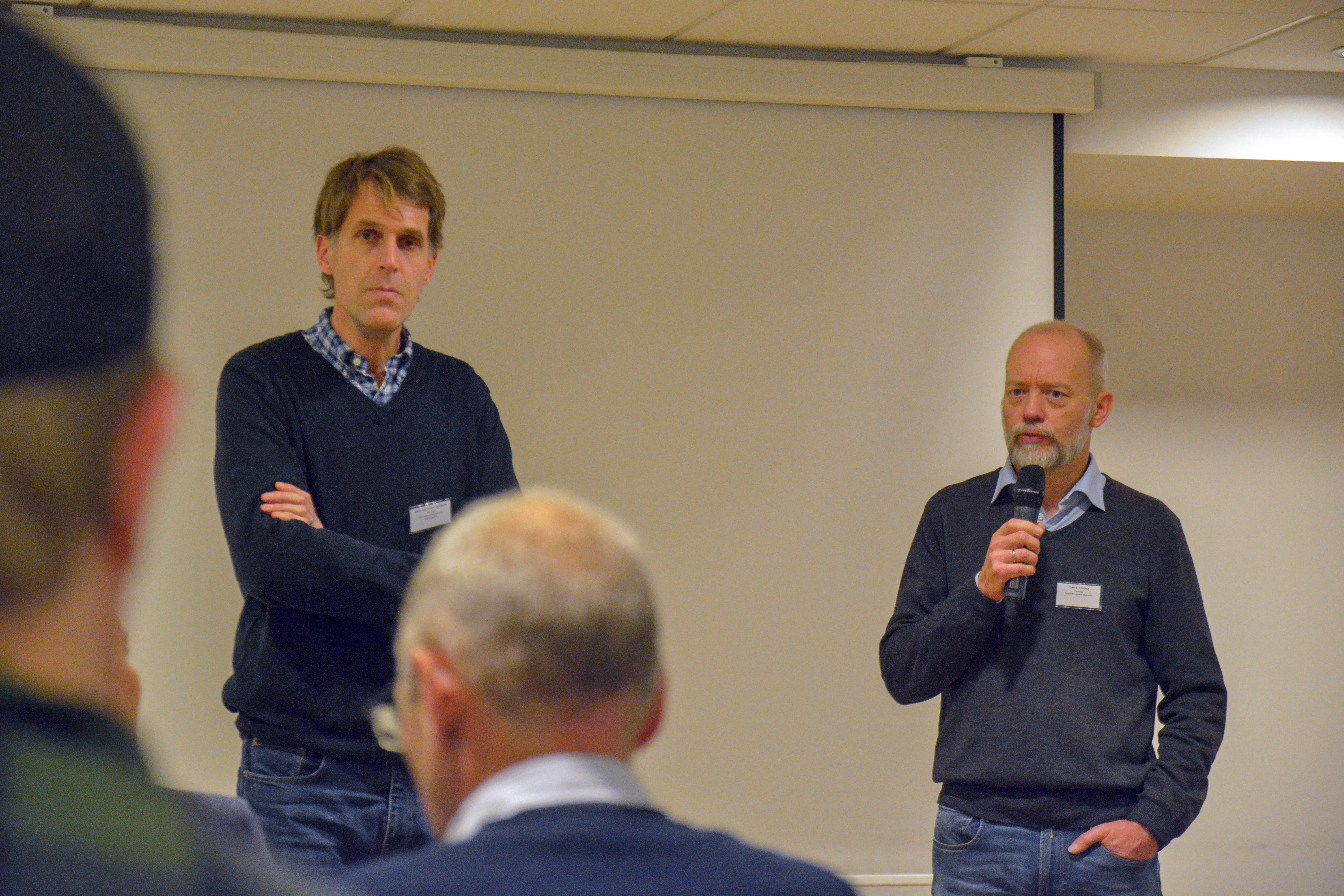 To menn står på en scene og holder foredrag. Begge mennene har på blå genser og står foran en hvit bakgrunn.