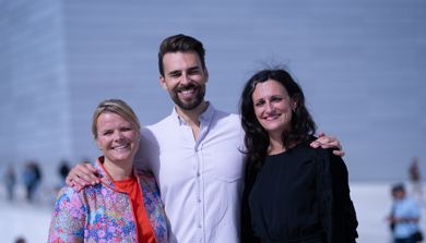 To kvinner og en mann står på operaen, arm-i-arm, og smiler mot kameraet. De får sola i øynene.