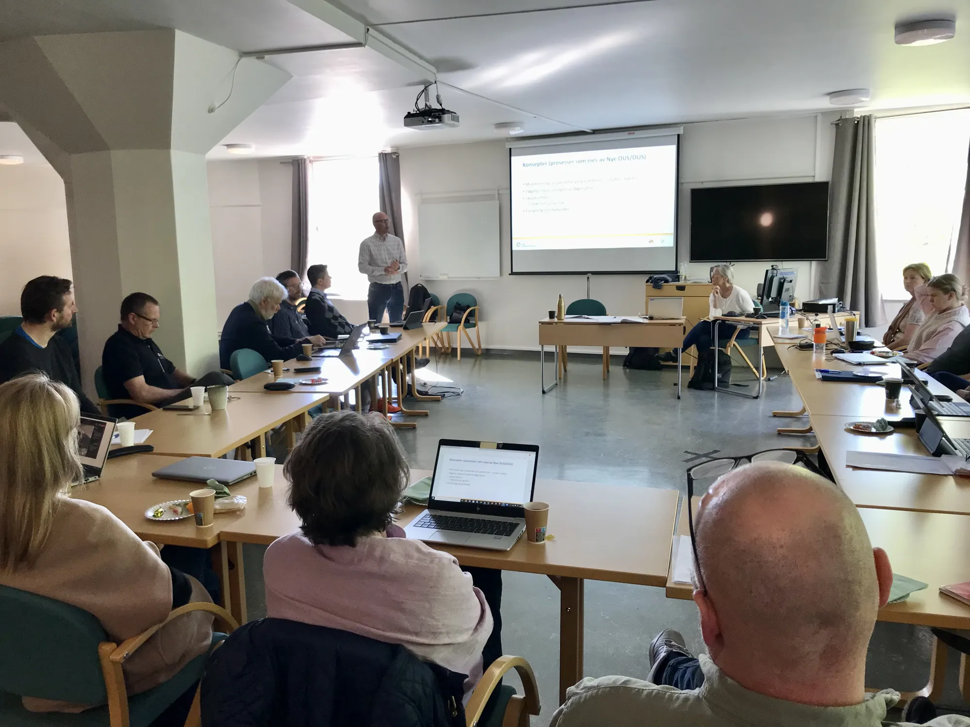 Bildet viser mange mennesker som sitter rundt bord og følger med på en presentasjon.
