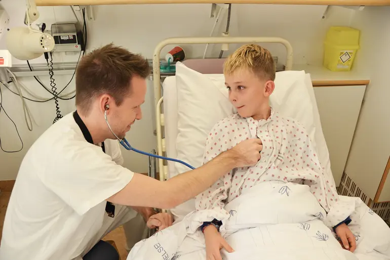En lege lytter på hjertet til en gutt som ligger i en sykehusseng