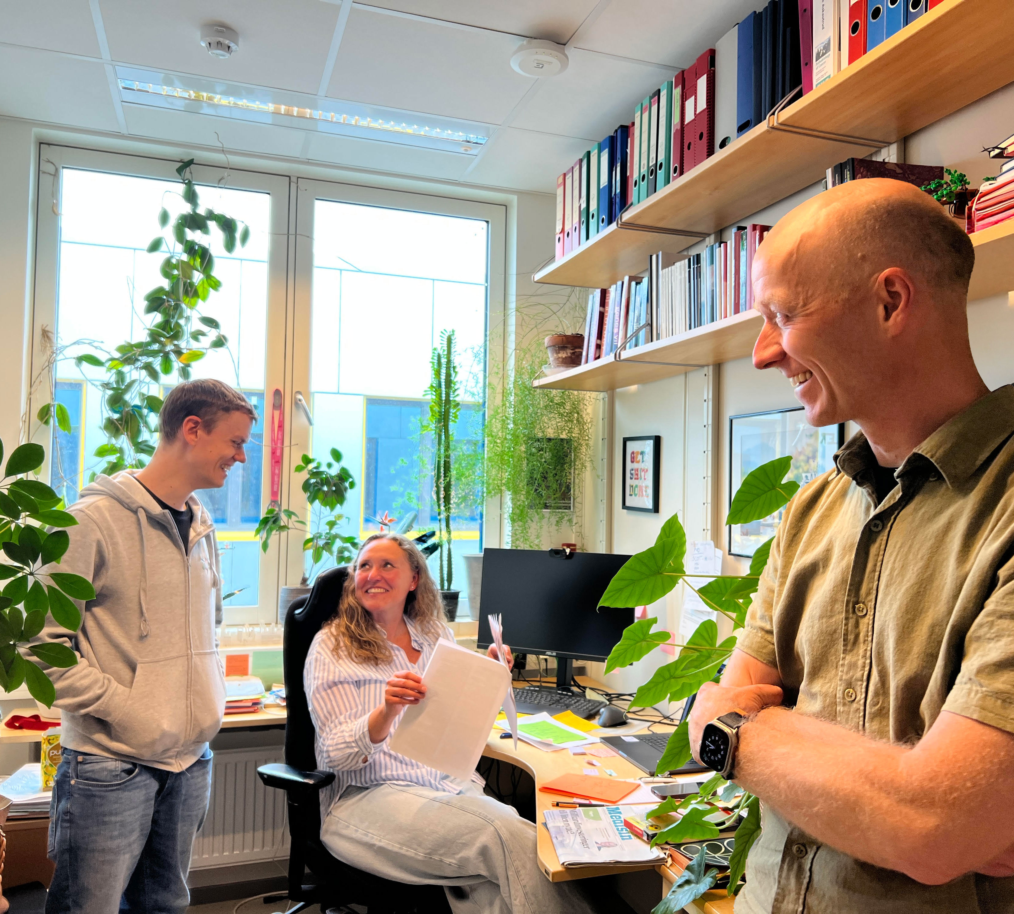 Image showing Andreas Kleppe, Hanne Askautrud and Tarjei Hveem in Hanne's office, when Andreas reveals the good news about funding from the Norwegian Research Council