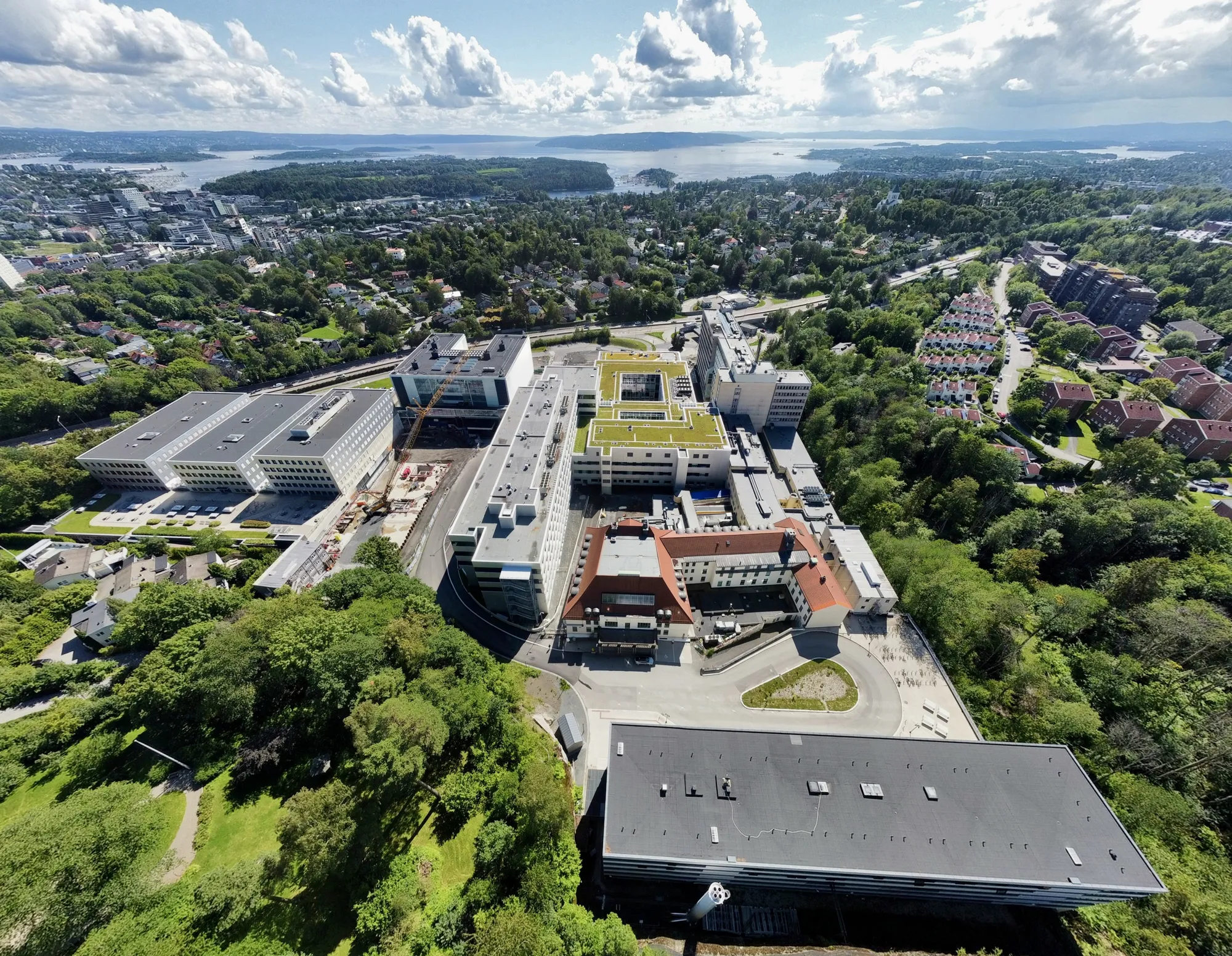 Radiumhospitalet sett fra oven blant grønne trær og med utsikt helt ned til Oslofjorden.