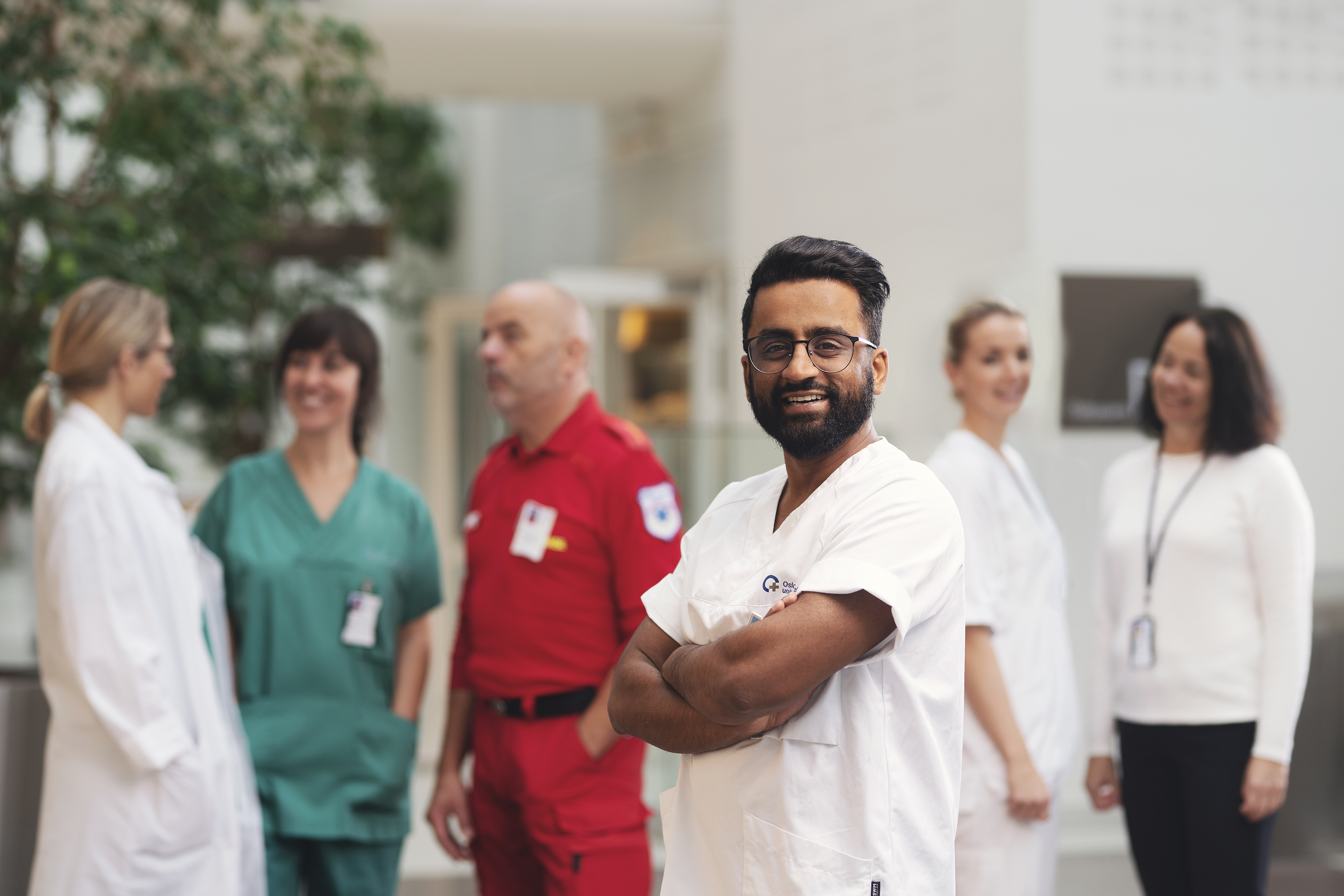 Seks ansatte ved OUS med ulike fagområder står sammen og prater. En av dem, med hvit t-skjorte, står foran i fokus og smiler mot kameraet.